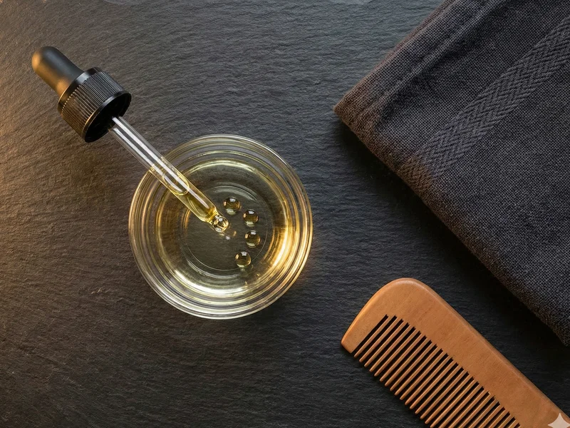 Dosage huile à barbe : pipette déposant des gouttes dorées dans une coupelle en verre