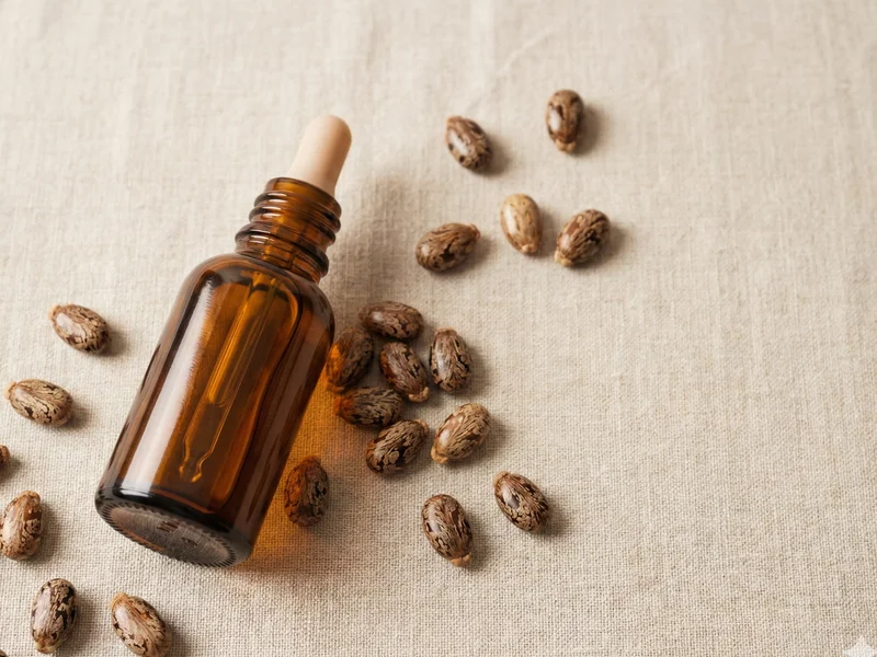 Flacon compte-gouttes d'huile de ricin ambrée posé sur un tissu en lin beige entouré de graines de ricin